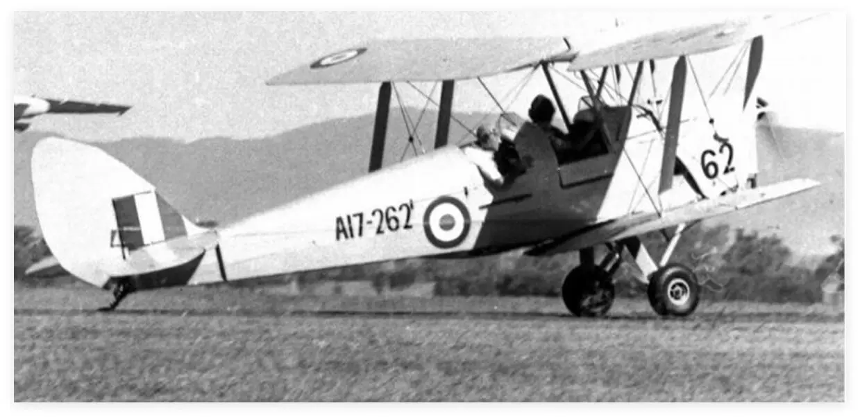 A historic black and white image of two Tiger Moth biplanes, including VH-IVN, parked near a hangar on a grassy airfield, capturing early aviation heritage A historic black and white image of two Tiger Moth biplanes, including VH-IVN, parked near a hangar on a grassy airfield, capturing early aviation heritage
