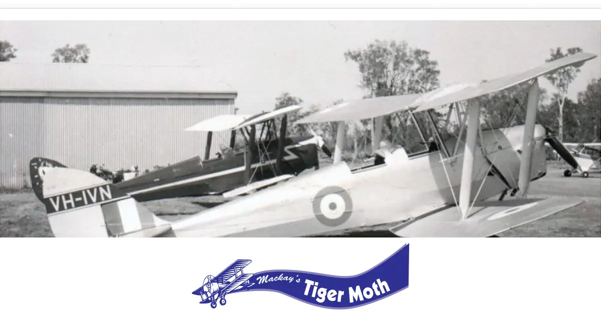 A historic black and white image of two Tiger Moth biplanes, including VH-IVN, parked near a hangar on a grassy airfield, capturing early aviation heritage