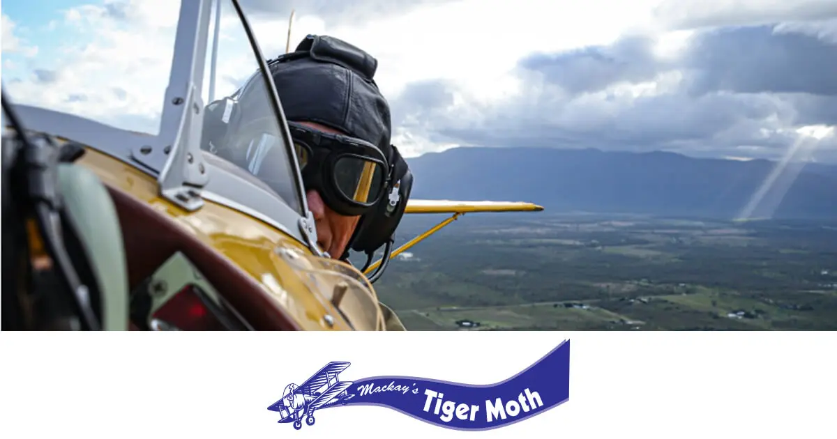 A pilot wearing vintage aviation goggles and a leather helmet looks back from the open cockpit of a Tiger Moth, with scenic countryside below.