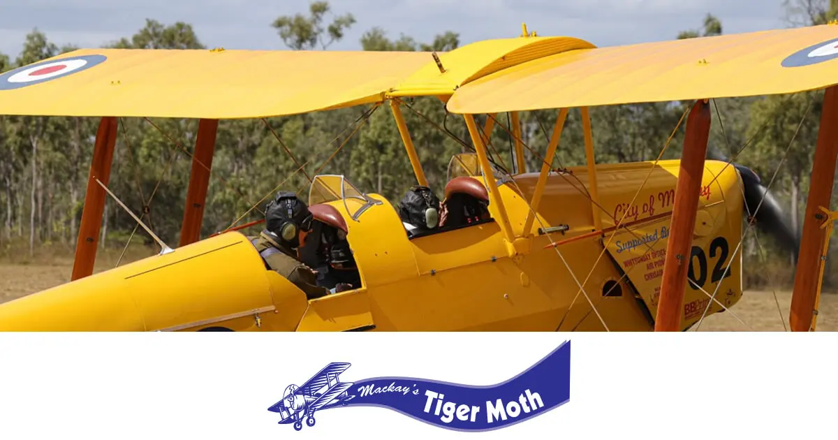 A vintage yellow Tiger Moth biplane, 'City of Mackay' (A17-302), with two pilots in the open cockpit, sits on a grassy airfield. Preserved by the Mackay Tiger Moth Museum.