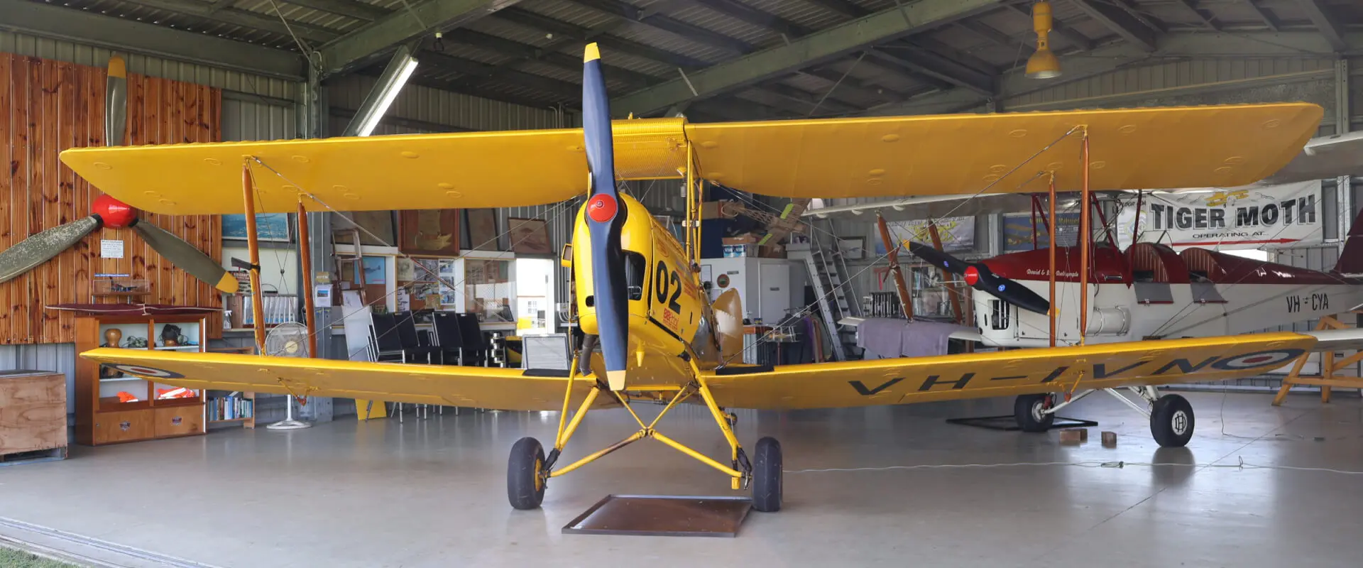 A vintage yellow Tiger Moth biplane, ‘City of Mackay’ (A17-302), with two pilots in the open cockpit, sits on a grassy airfield. Preserved by the Mackay Tiger Moth Museum. A vintage yellow Tiger Moth biplane, 'City of Mackay' (A17-302), with two pilots in the open cockpit, sits on a grassy airfield. Preserved by the Mackay Tiger Moth Museum.