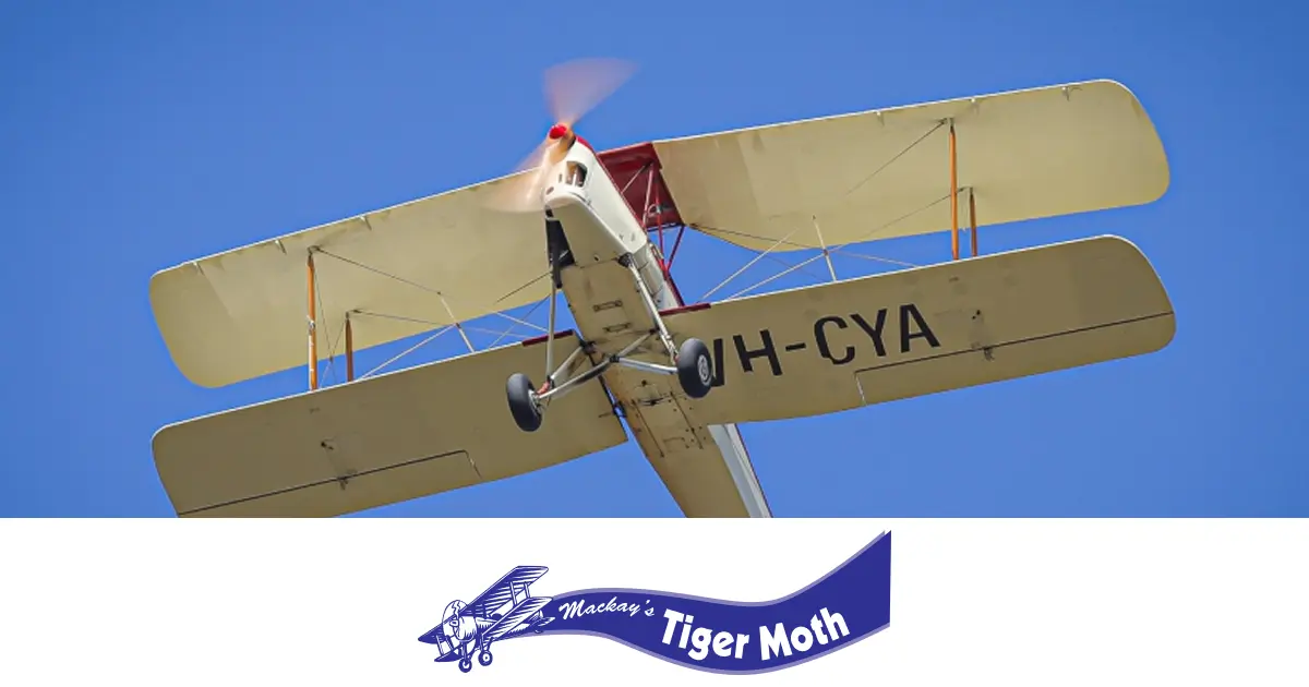 A white and red Tiger Moth biplane, registration VH-CYA, soars against a clear blue sky with its propeller spinning