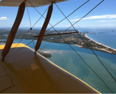A Tiger Moth biplane flying above a coastal landscape, with turquoise waters and sandy shores stretching towards the horizon A Tiger Moth biplane flying above a coastal landscape, with turquoise waters and sandy shores stretching towards the horizon