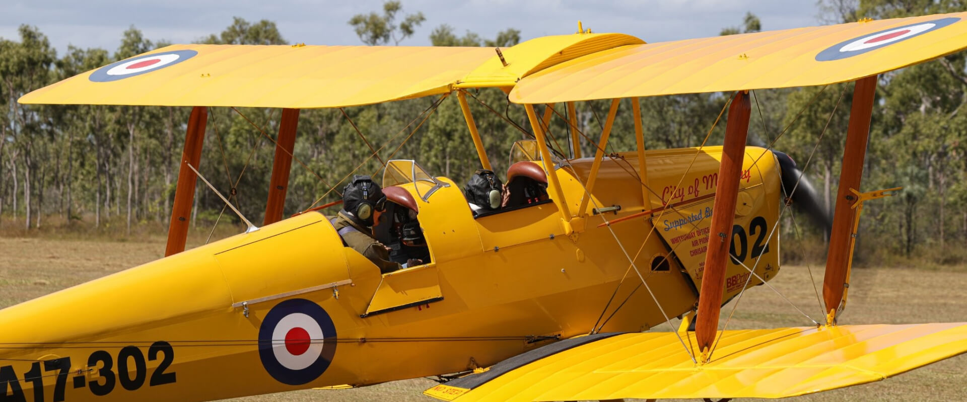 A vintage yellow Tiger Moth biplane, 'City of Mackay' (A17-302), with two pilots in the open cockpit, sits on a grassy airfield. Preserved by the Mackay Tiger Moth Museum.