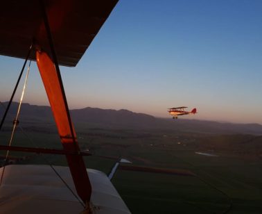 The view from the cockpit of a Tiger Moth biplane in flight, looking over lush green fields and distant mountain ranges under a partly cloudy sky