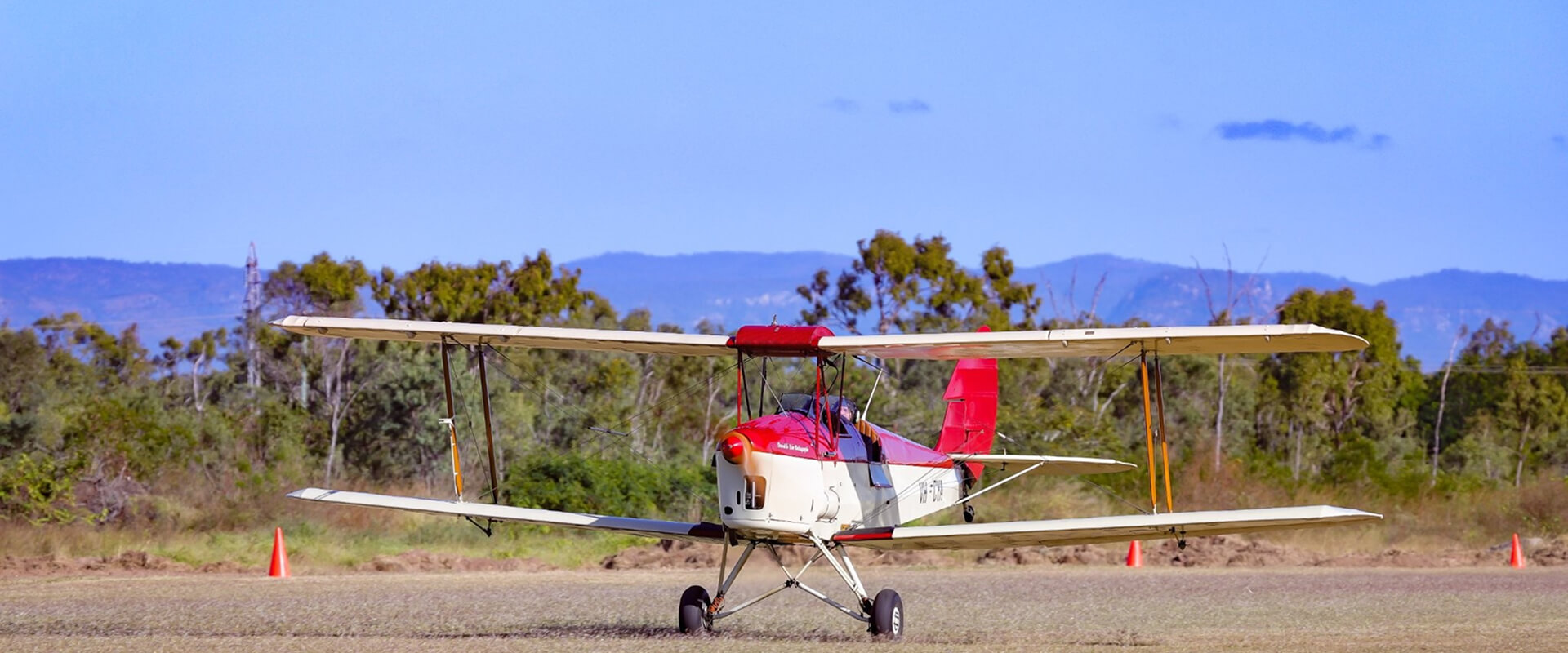 A red and white de Havilland Tiger Moth biplane taxiing on a grassy airstrip, with trees, mountains, and traffic cones in the background. The open cockpit reveals the pilot preparing for takeoff.