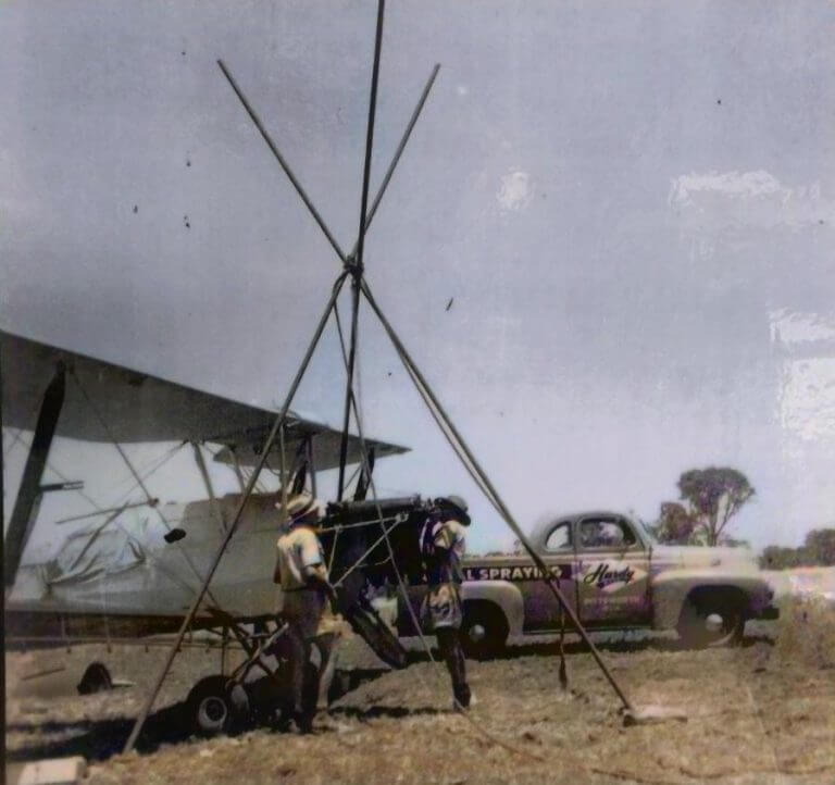 A pilot wearing goggles and a thick coat sits in the open cockpit of a Tiger Moth aircraft modified for aerial spraying, with a chemical spray tank visible behind the seat.