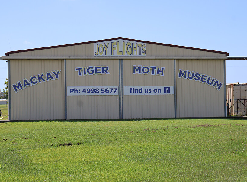 A large beige hangar with "Mackay Tiger Moth Museum" and "Joy Flights" written in blue and yellow letters. A large beige hangar with "Mackay Tiger Moth Museum" and "Joy Flights" written in blue and yellow letters.