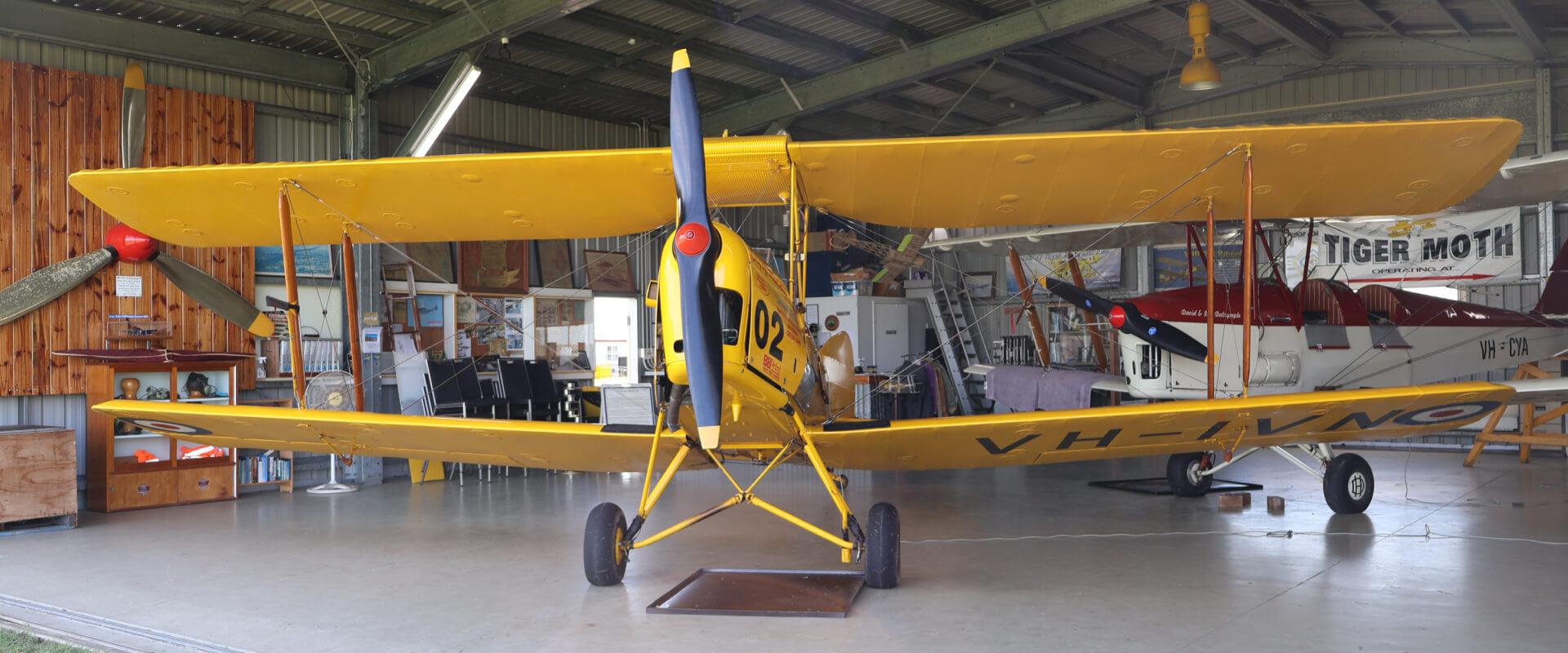 A vintage yellow Tiger Moth biplane, 'City of Mackay' (A17-302), with two pilots in the open cockpit, sits on a grassy airfield. Preserved by the Mackay Tiger Moth Museum.